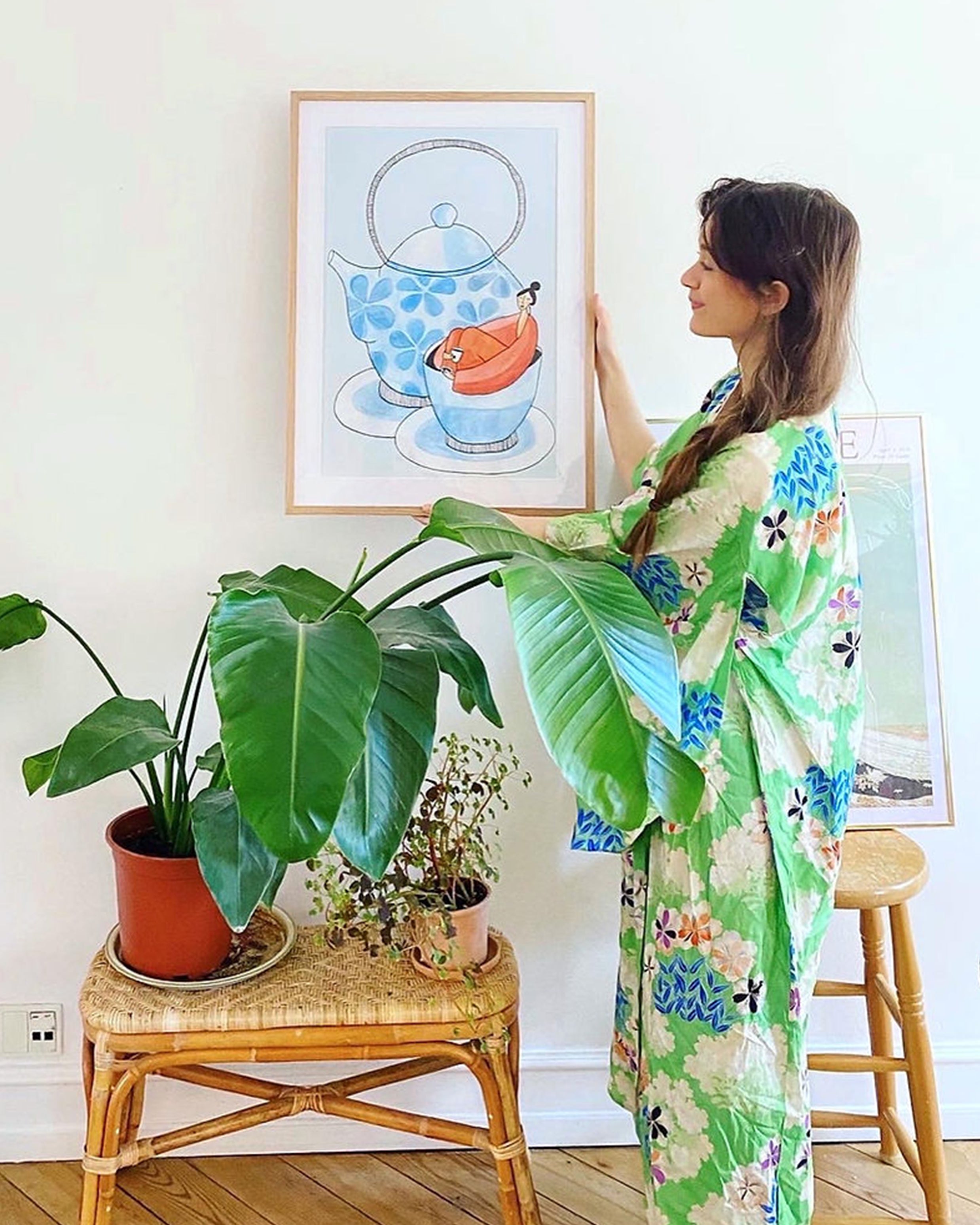 Kvinde i grøn blomstret kimono holder ROOIBOS kunstplakat i naturtræsramme ved grønne planter